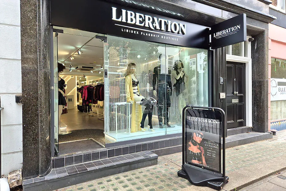 The Liberation shopfront at 4 Percy Street: an illuminated white LIBERATION wordmark on a black fascia above an open glass frontage, with mannequins in yellow and black latex in the window and a pavement sign reading Latex Clothing & Accessories.