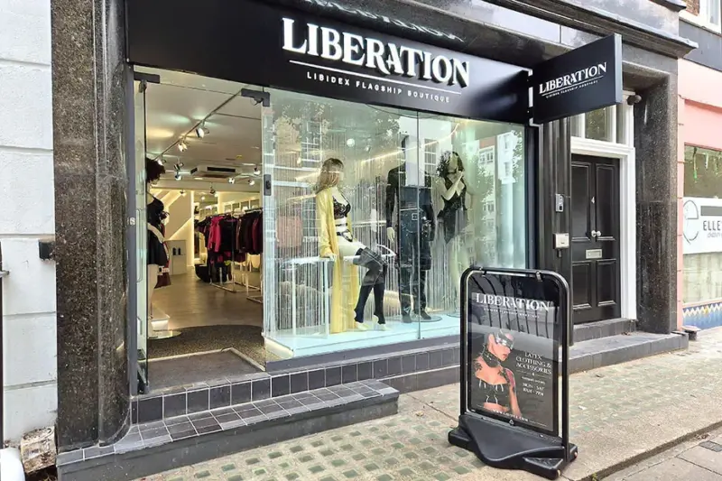 The Liberation shopfront at 4 Percy Street: an illuminated white LIBERATION wordmark on a black fascia above an open glass frontage, with mannequins in yellow and black latex in the window and a pavement sign reading Latex Clothing & Accessories.