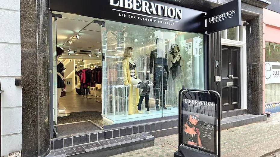 The Liberation shopfront at 4 Percy Street: an illuminated white LIBERATION wordmark on a black fascia above an open glass frontage, with mannequins in yellow and black latex in the window and a pavement sign reading Latex Clothing & Accessories.